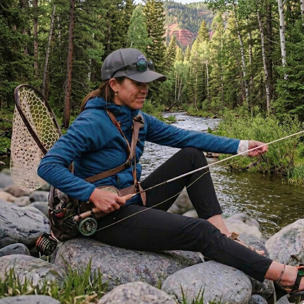 Femme pêchant à la mouche assise sur des rochers au bord d'une rivière de montagne entourée de forêt automnale