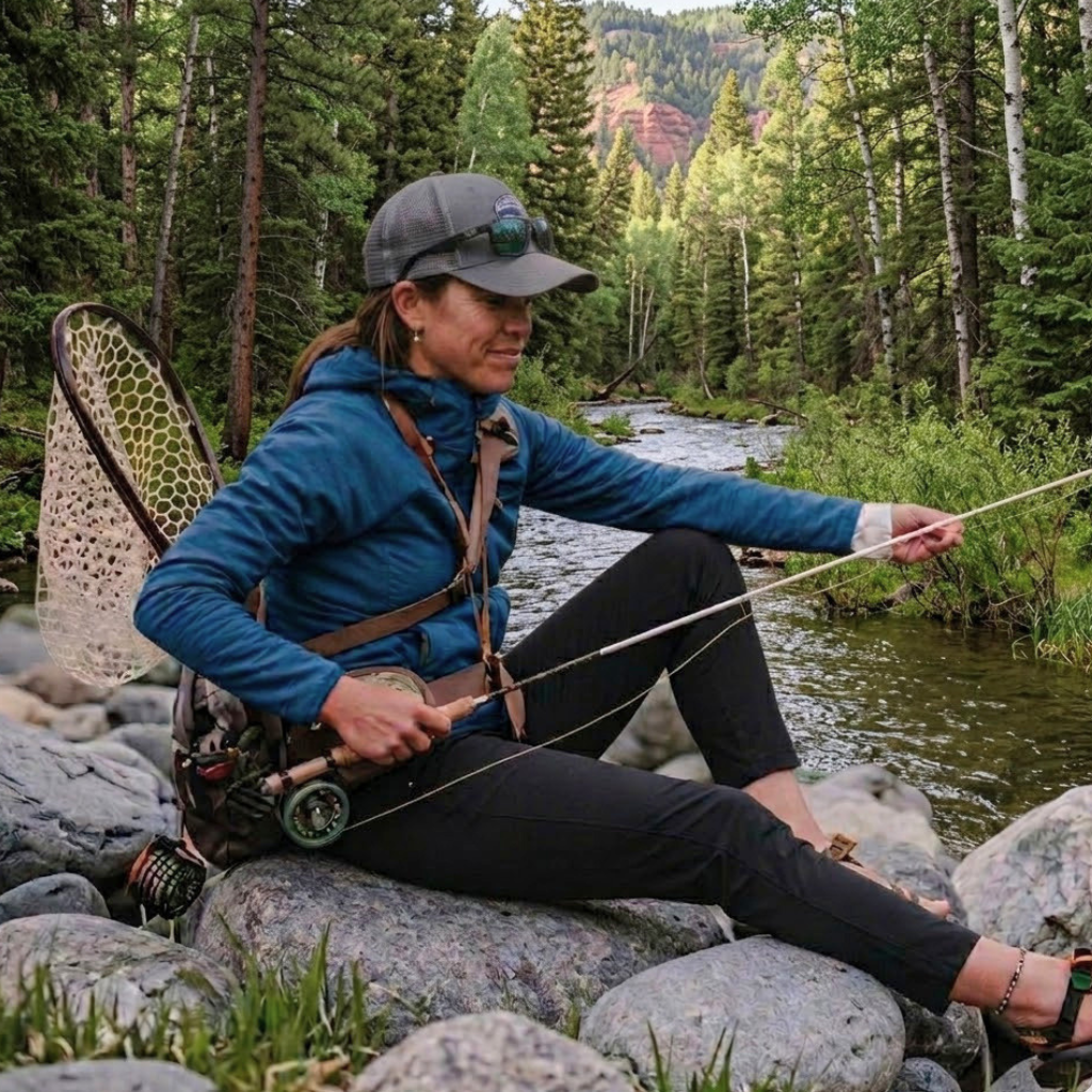 Femme pêchant à la mouche assise sur des rochers au bord d'une rivière de montagne entourée de forêt automnale