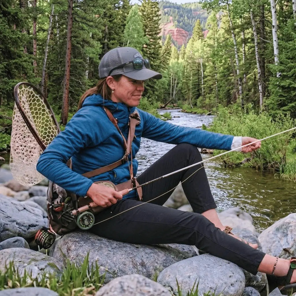 Femme au bord d'une rivière ajustant sa canne à mouche, représentant l'esprit technique et la connexion avec la nature de la pêche à la mouche.
